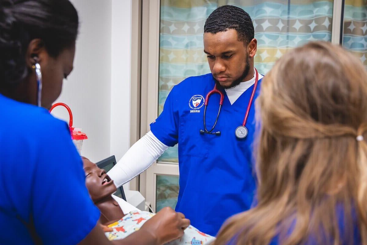 A nursing student in blue scrubs works on a manikin in a simulation lab